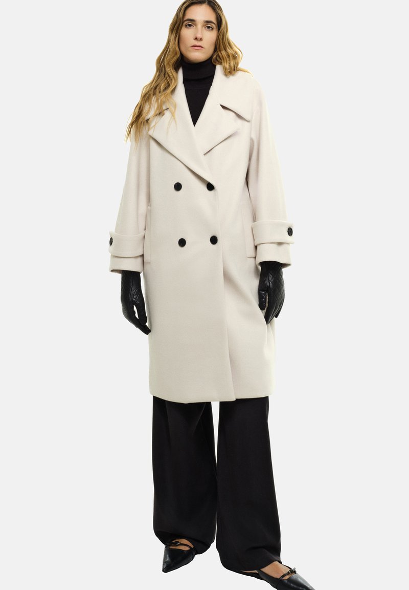 Cremefarbener Peacoat mit doppelt geknöpftem Vorn, schwarzen Knöpfen und breiten Manschetten. Das Model trägt einen schwarzen Rollkragenpullover und weite schwarze Hosen und hält schwarze Handschuhe in der Hand.