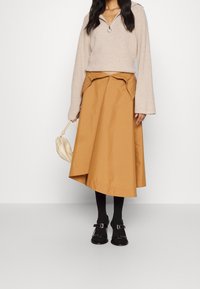 Beiger Strickpullover mit Reißverschlusskragen, kombiniert mit einem braunen A-Linien-Rock, schwarzen Strumpfhosen und schwarzen Schnallenschuhen, hält eine hellfarbene Handtasche.