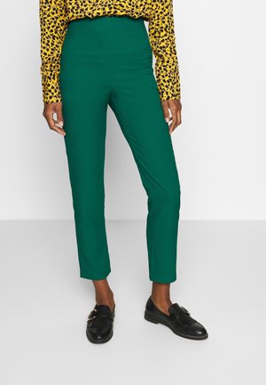 Person, die hochgeschnittene grüne, schmal zulaufende Hose, ein gelbes Langarmshirt mit Leopardenmuster und schwarze Loafers trägt und auf einem weißen Boden steht.