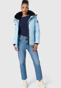 Lichtblauwe pufferjack met een zwart nepbont kraag, ritssluiting en contrasterende stiksels, gecombineerd met blauwe jeans en witte sneakers.