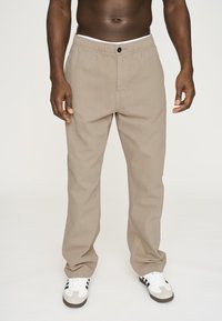 Beige bukser lavet af bomuld med en afslappet pasform. Har en elastisk talje og en enkelt knaplukning. Båret med hvide og sorte sneakers.