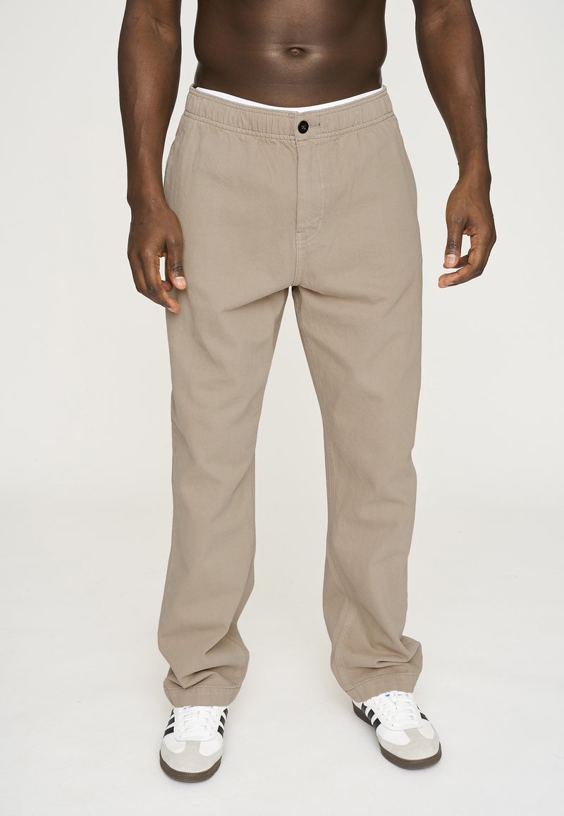 Beige bukser lavet af bomuld med en afslappet pasform. Har en elastisk talje og en enkelt knaplukning. Båret med hvide og sorte sneakers.