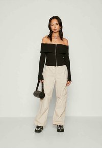 Schwarzes geripptes Off-Shoulder-Top mit Reißverschluss, cremefarbene Cargo-Hosen, schwarze runde Handtasche und schwarze Sneakers mit weißen Akzenten.