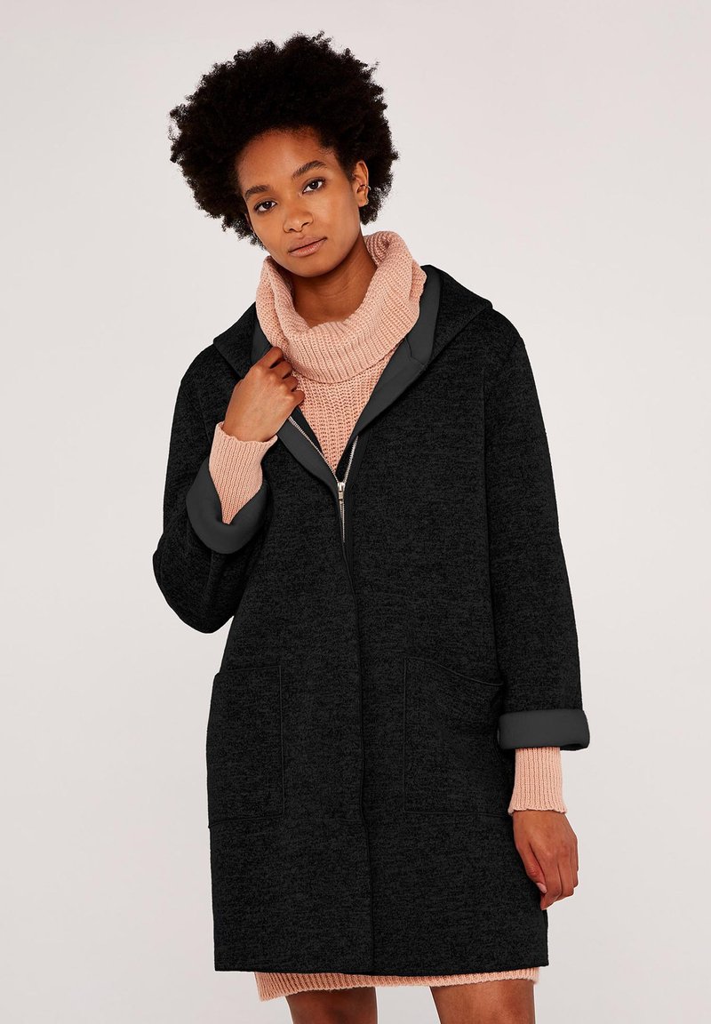 Svart huva coat med ett mjukt, texturerat tyg, med dragkedja fram, två fickor och upplindade ärmar, bärs över en rosa stickad tröja.