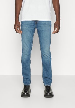 Homme portant un jean slim bleu clair, des chaussures noires à lacets et une chemise blanche, debout devant un fond uni.