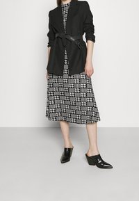 Blazer noir avec une ceinture nouée, sur une robe à motif pied-de-poule noir et blanc. Mules pointues noires. Texture lisse, coupe ajustée.