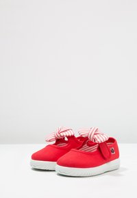 Zapatos rojos para niño pequeño con suelas de goma blancas y lazos de tela a rayas rosas y blancas encima, colocados sobre una superficie blanca.