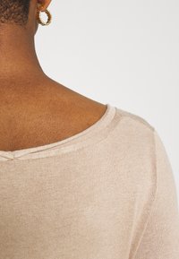 Pull beige avec une texture douce et côtelée, doté d'un large col rond et de détails subtils de couture aux épaules.
