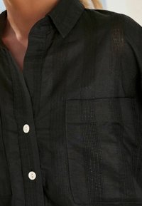 Nærbillede af en sort button-up skjorte med subtil vertikal stribet tekstur og en brystlomme, båret af en person med synlig hals og kraveben.