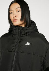 Melna pūkainā jaka ar kapuci, ar priekšējo rāvējslēdzēju un regulējamiem aukliņām. Iekļauts balts Nike logo uz krūtīm.