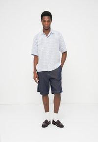 Μπλε καρό πουκάμισο με κοντά μανίκια και κουμπιά, συνδυασμένο με σκούρο τζιν σορτς και μαύρα παπούτσια, συμπληρωμένο με λευκές κάλτσες.