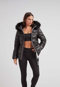 Czarna kurtka puchowa z kapturem z sztucznego futra, zasuwana z przodu, z bocznymi kieszeniami. Ozdobiona paskami przy zamku błyskawicznym oraz etykietą logo po boku.