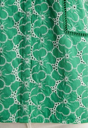 Zöld hímzett szövet, kör alakú mintákkal és virágos díszítésekkel, fehér varrási részletekkel és textúrázott felülettel.