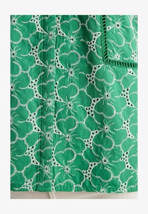 Grøn broderet stof med cirkulære mønstre og blomsteraccenter, med hvide søm detaljer og en struktureret overflade.