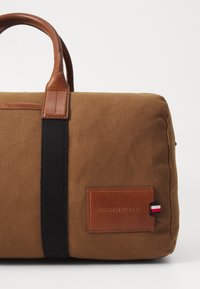 Borsa da viaggio in tela marrone con manici in pelle beige, una tracolla nera e una targhetta in pelle con un piccolo accento tricolore e logo.