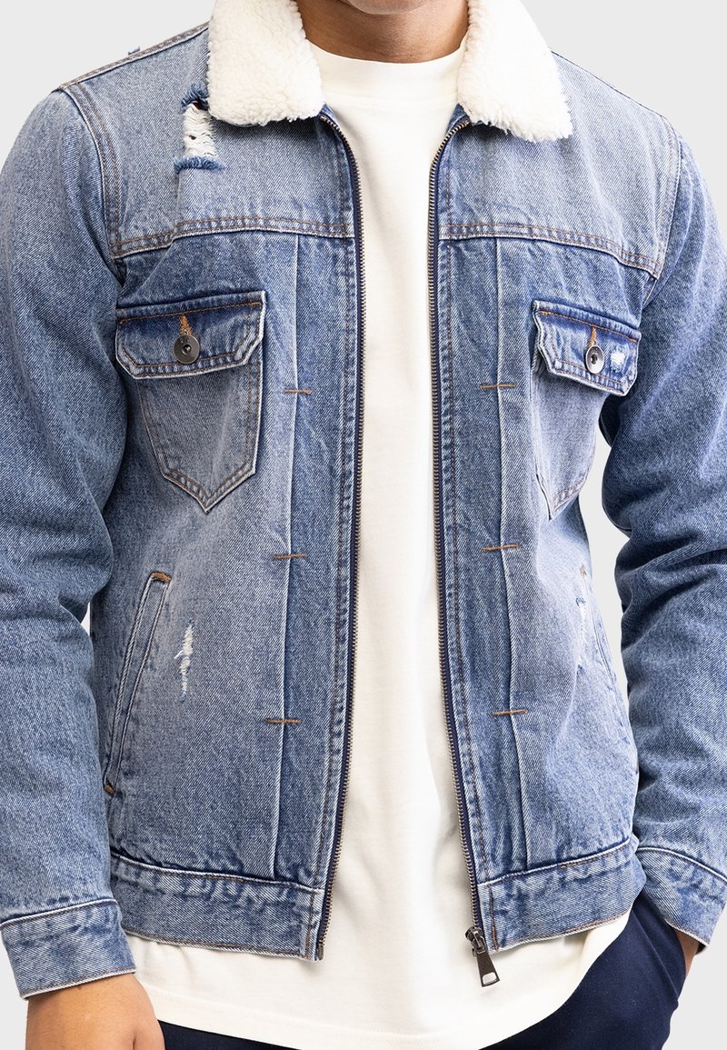Kurtka dżinsowa z kołnierzem z futerka typu shearling, jasnoniebieskiego koloru, z przetarciami, zamkiem błyskawicznym z przodu, dwoma kieszeniami na piersiach i brązowymi przeszyciami jako akcentami.
