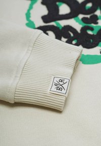 Kermanvärinen huppari, jossa on raidalliset hihansuut, brodeerattu musta teksti ja vihreä yksityiskohta. Tagi esittelee mustavalkoista logoa.