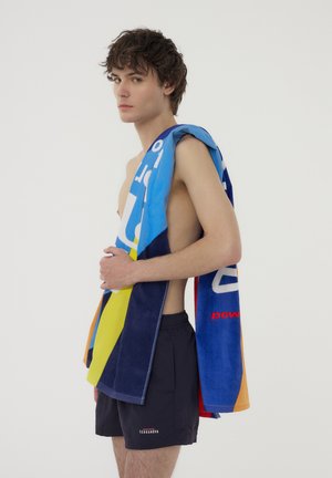 Asciugamano di spugna multicolore con motivi geometrici, prevalentemente blu, giallo e blu scuro, drappeggiato su una spalla nuda. Indossato con pantaloncini da bagno neri.