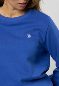 Felpa di cotone blu con scollatura a girocollo, caratterizzata da un piccolo logo ricamato bianco sul petto. Texture morbida e maniche lunghe.