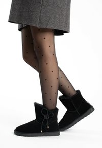 Botas de tobillo de ante negro con un suave forro de piel, que presentan un detalle de lazo atado y suelas planas de goma, llevadas con medias negras con puntos.