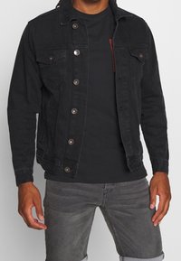 Chaqueta de mezclilla negra con cierre de botones, dos bolsillos en el pecho y botones metálicos en contraste. Llevada sobre una camiseta negra, combinada con jeans grises.