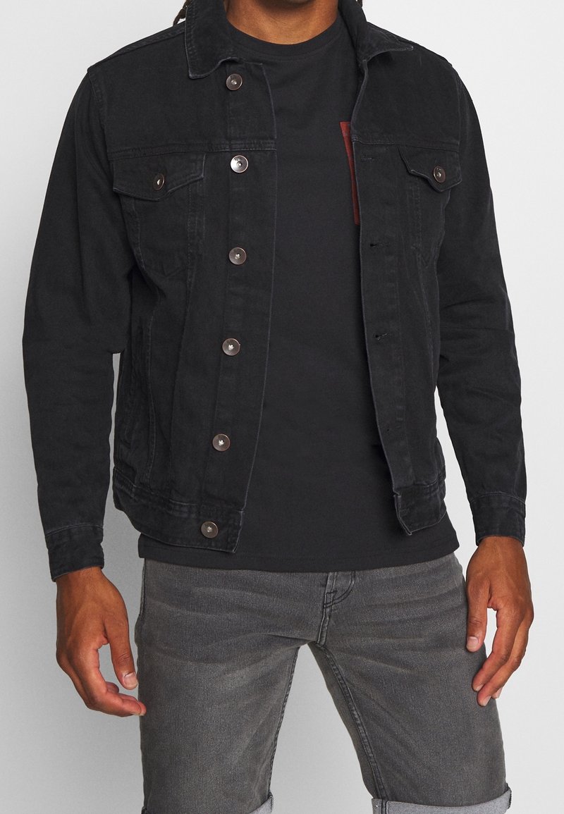 Chaqueta de mezclilla negra con cierre de botones, dos bolsillos en el pecho y botones metálicos en contraste. Llevada sobre una camiseta negra, combinada con jeans grises.
