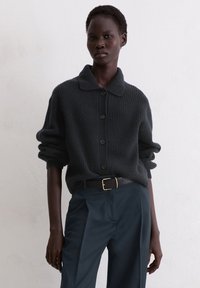 Cardigan en maille côtelée gris foncé avec col à revers et boutons, associé à un pantalon sombre et une ceinture noire avec boucle dorée.