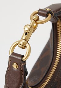 Gros plan sur une sangle de sac en cuir marron avec fermoir métallique doré et anneaux, dents de la fermeture éclair visibles et marquage "Coach" sur la quincaillerie.