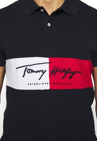 Polo navy con una franja horizontal blanca y roja que presenta texto negro. El texto dice "Tommy Hilfiger" y "ESTABLECIDO MCMLXXXV."