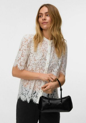 Femme en blouse blanche en dentelle à manches courtes, tenant un petit sac à main noir, regardant légèrement de côté avec des cheveux blonds lâches.