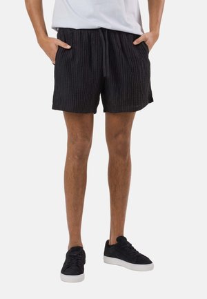 Person iført sorte, teksturerede shorts med lommer, hvid t-shirt og sorte sneakers med hvide såler, stående med hænderne i lommerne.