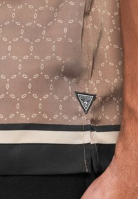 Hnedá košeľa so vzorom na leme s čiernymi a béžovými pruhmi a malou čierno-bielou nášivkou loga Guess pri zápästí osoby, spolu s čiernymi nohavicami.