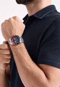 Reloj de acero inoxidable plateado con bisel rojo y azul, esfera negra y funciones de cronógrafo, llevado en una mano, combinado con una camisa oscura texturizada.