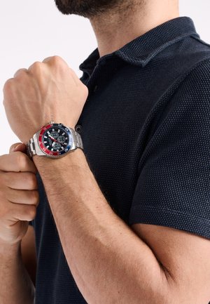 Montre en acier inoxydable argenté avec une lunette rouge et bleue, un cadran noir et des fonctions chronographe, portée sur une main, assortie à une chemise sombre texturée.