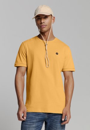 Geel katoenen T-shirt met een ronde hals en korte mouwen, voorzien van een klein donker logo op de borst. Gecombineerd met een beige pet.