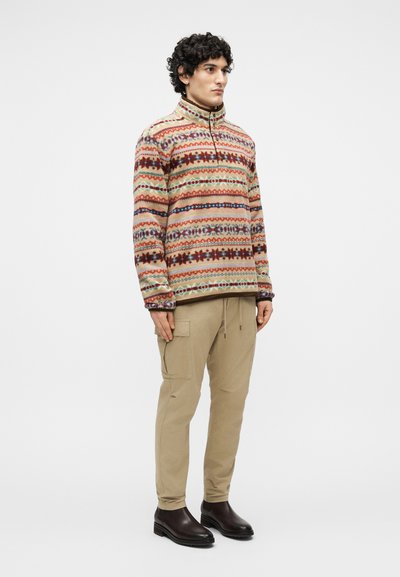 Mees, kes seisab beežides cargo-püksides, tumepruunides saapades ja mitmevärvilise mustriga fliisist poole lukuga pulloveris.