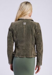 Een vrouw met lang blond haar draagt een groene leren jas met een schapenvachtkraag, gezien vanuit de rug, waarop de details van de jas goed te zien zijn.