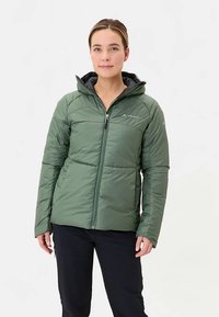 Groene geïsoleerde jas met ritssluiting, capuchon en doorgestikt ontwerp. Voorzien van een logo en gedragen over een donkere broek. Textuur van de stof is zichtbaar.