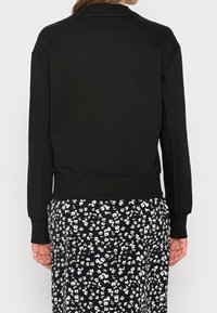 Sudadera negra con cuello y puños de canalé, con un corte holgado, mostrada desde la parte posterior con una falda de estampado floral.