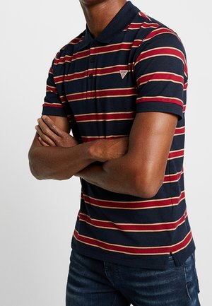 Mand med foldede arme iført en navy blå polo med røde og beige vandrette striber og mørkeblå jeans.