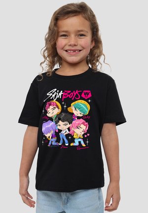 Lächendes Kind trägt ein schwarzes T-Shirt mit bunten Cartoon-Figuren, Namen und dem Text "Saja Boys" auf der Vorderseite.