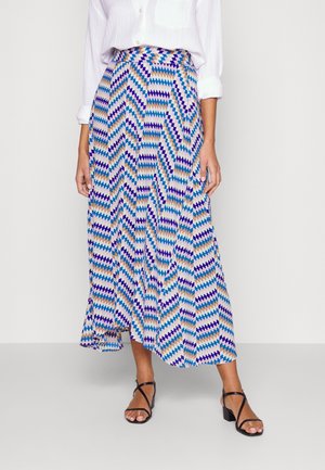 Femme portant une chemise blanche à manches longues rentrée dans une jupe plissée taille haute aux motifs zigzag bleu, beige et violet, associée à des talons noirs à brides.