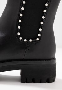 Bota de tobillo de cuero negro con panel lateral elástico delineado por tachuelas plateadas y un tacón de suela robusta.