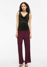 Top senza maniche in satin nero con drappeggio frontale, abbinato a pantaloni larghi plissettati borgogna. L'insieme è completato da scarpe con tacco aperto nere.