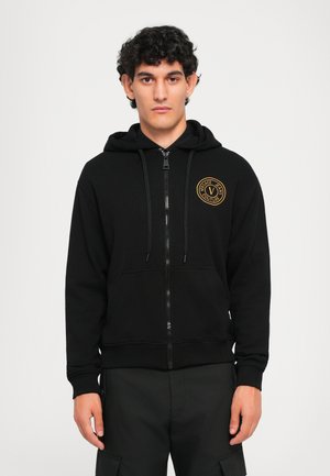 Ung mann med krøllete svart hår iført svart zip-up hettegenser med gull sirkulært logo på brystet, stående mot en enkel lys bakgrunn.