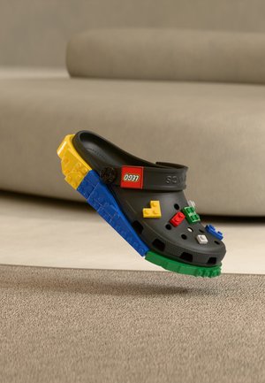 Μαύρο παπούτσι Crocs διακοσμημένο με πολύχρωμα τούβλα LEGO στη σόλα και το επάνω μέρος, αιωρούμενο πάνω από μπεζ χαλί.