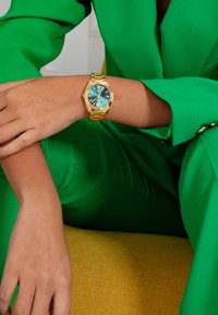 Montre-bracelet en or avec un cadran teal, des marqueurs en chiffres romains et un bracelet à maillons en or, posée sur une surface beige clair et jaune.