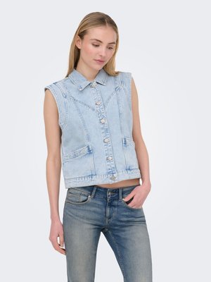 Jonge vrouw draagt een lichtblauw denim mouwloos vest met knopen en een blauwe spijkerbroek, staand met één hand in de zak.