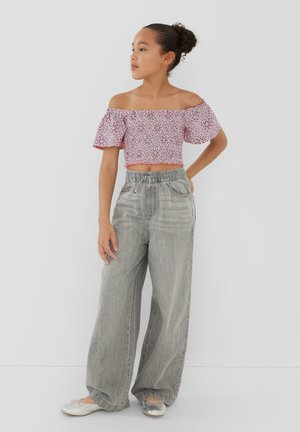 Meisje met haar in een knot, draagt een roze bloemen off-shoulder crop top, losse grijze broek en zilveren ballerina's, staande met hand op heup.