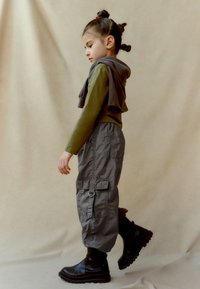 Top verde cu maneci lungi, cu un hanorac gri aruncat pe umeri, pantaloni cargo gri cu buzunare și cizme negre cu toc gros, toate pe un fundal neutru.